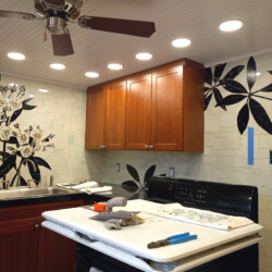 A kitchen with wooden cabinets, floral tile mural on the wall, and tools on the stove indicating ongoing renovations.