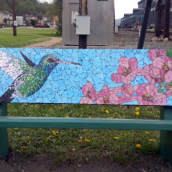 A mosaic bench backrest depicts a hummingbird near pink flowers against a blue background in an outdoor setting.
