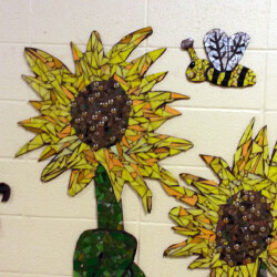 Mosaic artwork of two sunflowers, a bee, and a ladybug displayed on a cream-colored tiled wall.