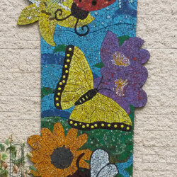 Colorful mosaic art panel featuring a ladybug, butterfly, bee, and flowers mounted on a light brick wall.