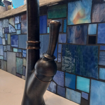 SOLD Close-up of a dark faucet mounted on a countertop with a blue mosaic tile backsplash in the background.
