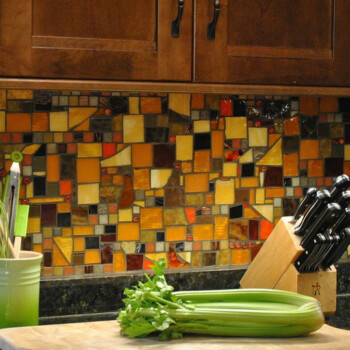 SOLD A cutting board with celery sits on a kitchen counter; knives and utensils are nearby, with a colorful mosaic backsplash.