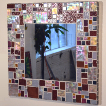 Square mirror with a mosaic frame of red, purple, and iridescent glass tiles, reflecting a window and plant.