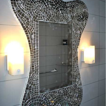 Rectangular mirror with an ornate, mosaic tile border, mounted on a tiled wall between two lit wall sconces.