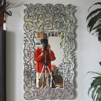Person in a red sweater taking a photo with a camera on a tripod, reflected in a decorative mosaic mirror.