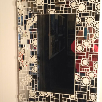 Rectangular mirror with a geometric, black and white mosaic tile frame, reflecting art pieces and part of a red object.