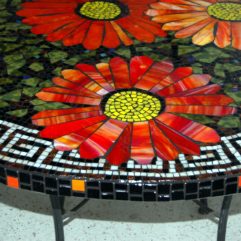 Round mosaic table featuring large red flowers with yellow centers and a black-and-white geometric border pattern.
