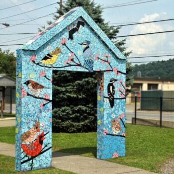 SOLD A mosaic archway decorated with colorful bird designs stands on a grassy area near a sidewalk and some buildings.