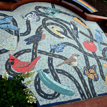 SOLD Colorful mosaic artwork depicting birds, butterflies, fruit, and winding paths set in a garden patio area.
