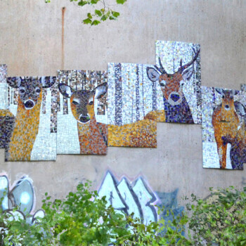 SOLD A mosaic mural of deer in a forest is mounted on a concrete wall above graffiti and surrounded by green foliage.