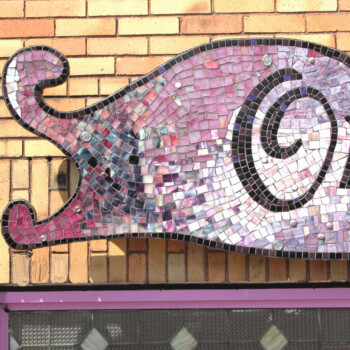 Pink and purple mosaic sign shaped like a cat on a yellow brick wall above a window with glass blocks.