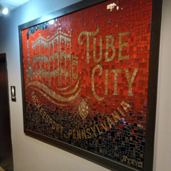 Mosaic wall art with "Tube City" and "Pennsylvania" in gold letters, located near a restroom sign.