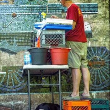 A man stands on crates, working on a mosaic wall, while a black dog lies underneath the table nearby.