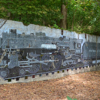 Mosaic mural of a steam locomotive and coal cars on a concrete wall in a wooded outdoor setting.