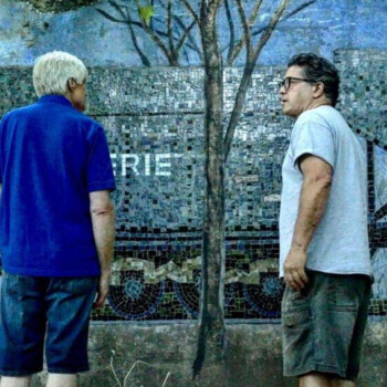 Two men stand in front of a mosaic mural depicting a train and trees; both are viewing the artwork.