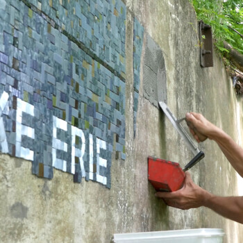 A person applies adhesive to a wall while assembling a mosaic mural that reads "LAKE ERIE" outdoors.